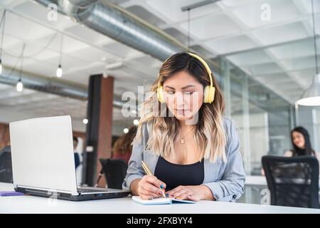 Geschäftsfrau mit Kopfhörern, die im Büro per Laptop Tagebuch schreibt Stockfoto