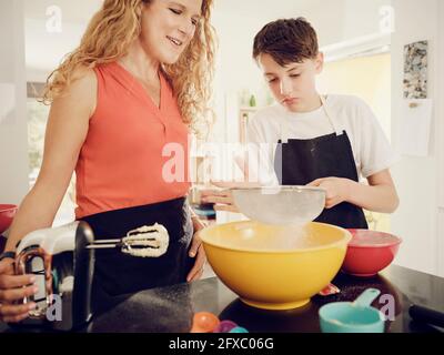 Mutter sah den Sohn an, der in der Schüssel Puderzucker sieben sah, während er in der Küche Essen zubereitete Stockfoto