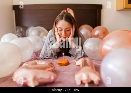 Frau mit dem Kopf in den Händen, die Kerze auf dem Donut ansah, während sie inmitten von Ballons lag Stockfoto