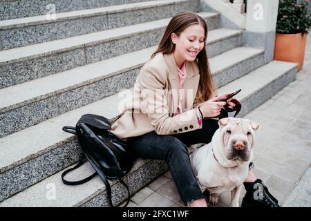 Schöne Frau mit Shar-Pei Hund mit Mobiltelefon, während sie auf der Treppe sitzt Stockfoto