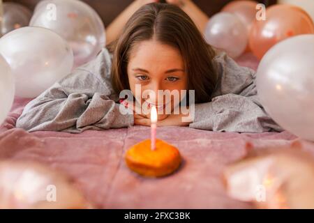 Junge Frau, die zu Hause auf dem Bett liegt und dabei die Kerze auf dem Donut brennt Stockfoto