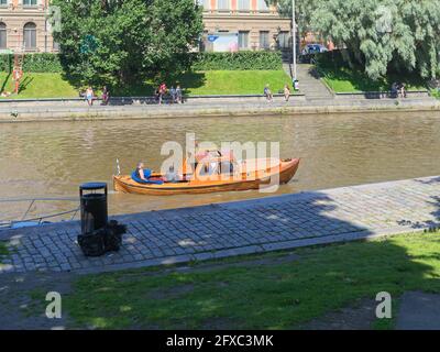 Die Menschen genießen einen sonnigen Sommertag am Ufer des Flusses Aura in Turku, Finnland, während ein Holzboot stromaufwärts fährt. Stockfoto