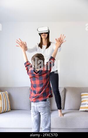 Lächelnde Mutter mit virtuellem Headset, die mit dem Sohn spielt, während sie im Wohnzimmer auf dem Sofa steht Stockfoto