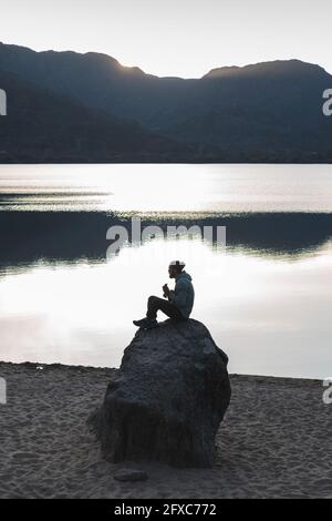 Mann, der während des Sonnenuntergangs auf einem Felsen am See sitzt Stockfoto