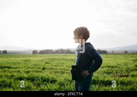 Netter Junge mit Fernglas, der wegschaut, während er an sonnigen Tagen auf dem Feld steht Stockfoto