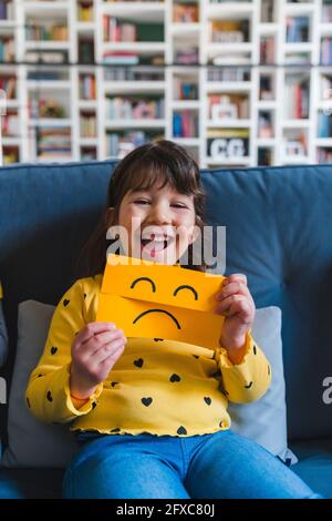 Lachendes Mädchen mit trauriger Emoticons im Gesicht, während es zu Hause auf dem Sofa sitzt Stockfoto