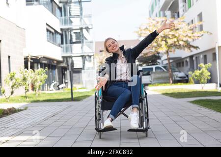 Glückliche behinderte Frau mit ausgestreckten Armen sitzt auf Rollstuhl in Wohngebiet Stockfoto