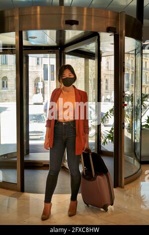 Frau in schützender Gesichtsmaske, die mit Koffer am Hoteleingang steht Stockfoto