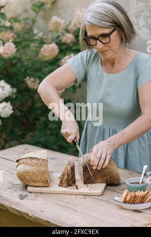 Reife Frau mit Brillen, die Brot auf dem Tisch schneiden Stockfoto