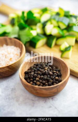 Holzschüssel mit schwarzen Pfefferkörnern mit gehackten Zucchinis im Hintergrund Stockfoto