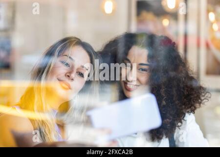 Glückliche Freundinnen, die Selfie auf dem Smartphone machen, gesehen durch das Glas im Restaurant Stockfoto