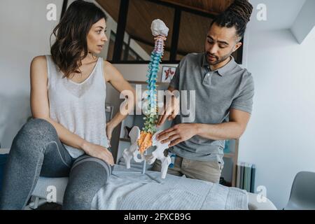 Männlicher Physiotherapeut erklärt weiblichen Patienten in der Praxis auf dem Massageliegen das Wirbelsäulenmodell Stockfoto