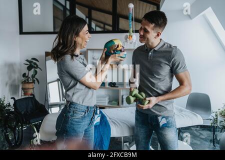 Fröhliche weibliche Physiotherapeuten diskutieren über Schädelmodell mit männlichen Kollegen mit Gewichten in der Praxis Stockfoto