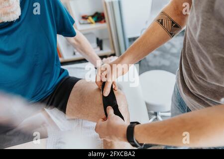 Männlicher Physiotherapeut klebt elastisches therapeutisches Band auf das Knie des Patienten Stockfoto