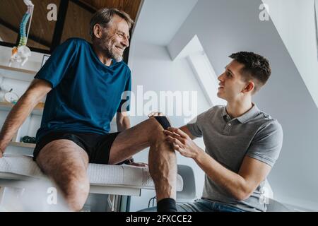 Männlicher Physiotherapeut klebt in der Praxis Kinesio-Klebeband auf das Knie des lächelnden Patienten Stockfoto