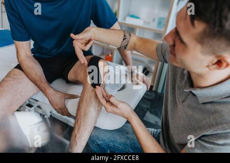 Der männliche Physiotherapeut klebt in der Praxis Kinesioband auf das Knie des Patienten am Massageliegen Stockfoto