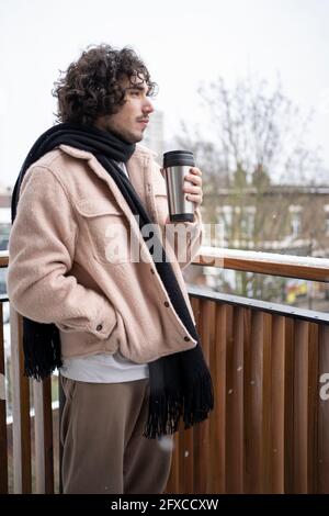 Nachdenklicher Mann in warmer Kleidung mit isoliertem Getränkebehälter auf dem Balkon Stockfoto