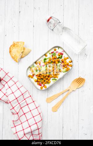 Studioaufnahme einer Flasche Wasser, Serviette, Holzbesteck, Fladenbrot und Lunchbox mit Joghurt mit Kichererbsen, Gurken und Radieschen Stockfoto