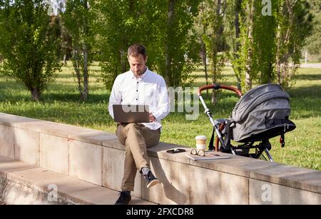 Ein mittelerwachsener Geschäftsmann, der am Laptop arbeitet, während er am Kinderwagen im Park sitzt Stockfoto