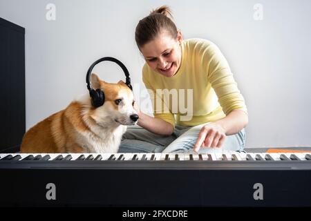 Lächelnde Frau mit Hund, der Kopfhörer trägt, während sie im Wohnzimmer Klavier übt Stockfoto