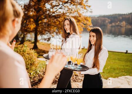 Die lächelnde Kellnerin hält an sonnigen Tagen ein Tablett mit Getränken am See Stockfoto