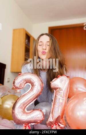 Junge Frau puckert, während sie die Nummer 21 Luftballons im Schlafzimmer zu Hause hält Stockfoto