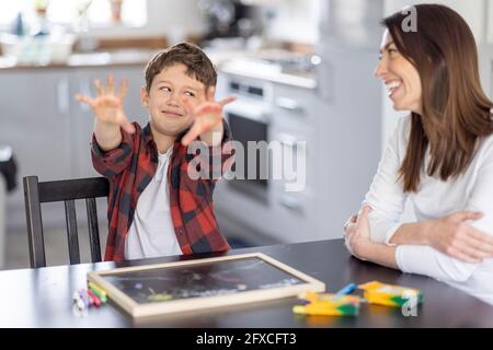 Junge, der schmutzige Hände zeigt, während er mit seitlichem Blick auf die Mutter in der Küche schaut Stockfoto