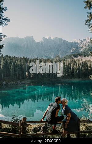 Zärtliches Paar küsst sich beim Sitzen auf dem Geländer am Karersee in Südtirol, Italien Stockfoto