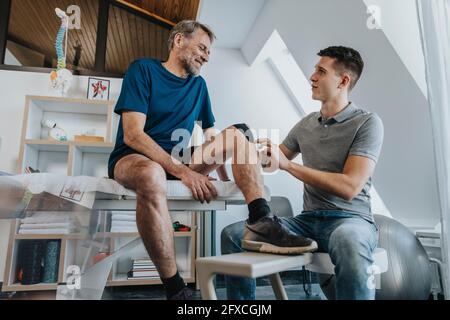 Männlicher Physiotherapeut, der lächelnde Patienten ansieht, während er in der Praxis elastisches therapeutisches Band klebt Stockfoto