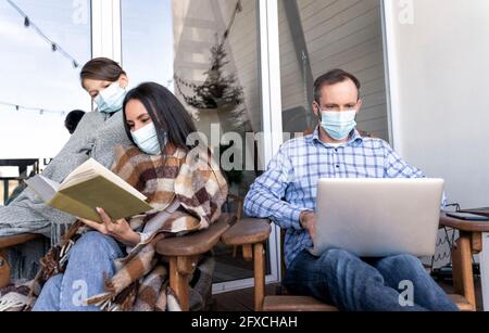 Mutter und Sohn lesen Buch, während der Vater während einer Pandemie einen Laptop auf dem Balkon benutzte Stockfoto