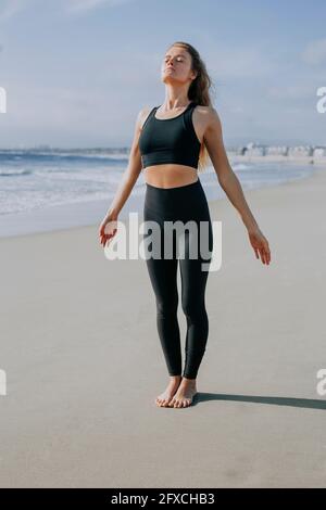 Junge Frau in Sportkleidung, die am sonnigen Tag mit geschlossenen Augen am Strand steht Stockfoto