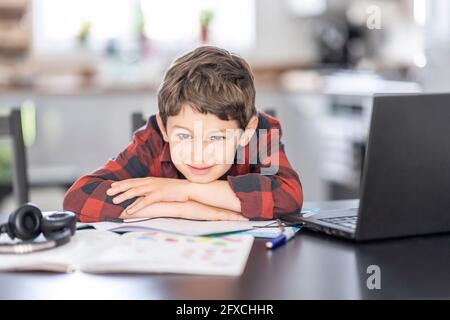 Lächelnder Junge, der sich zur Hand lehnt, während er zu Hause am Laptop sitzt Stockfoto