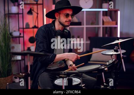 Bärtiger junger Mann mit schwarzem Hut und Sonnenbrille, der im Studio mit dunkler Atmosphäre elektronische Trommeln spielt. Konzept von Musik, Hobby und Inspiration. Stockfoto