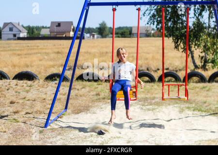 Unschärfe kleines Mädchen schwingen auf Schaukel auf Spielplatz mit. Ländliche Gegend. Leuchtend blaue und rote Schaukel. Kinder Sommerspiel. Mädchen haben Spaß im Sommer Stockfoto