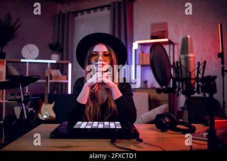 Porträt einer stilvollen jungen Frau in trendigem schwarzen Hut und Sonnenbrille, die auf die Kamera schaut, während sie mit verschiedenen Musikinstrumenten am Tisch sitzt. Musikerin im Heimstudio. Stockfoto