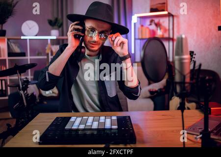 Bärtiger Mann mit schwarzem Hut und Brille, der Musik im Heimstudio mit DJ-Controller, Kopfhörern und Mikrofon aufnimmt. Konzept der Inspiration und kreatives Hobby. Stockfoto
