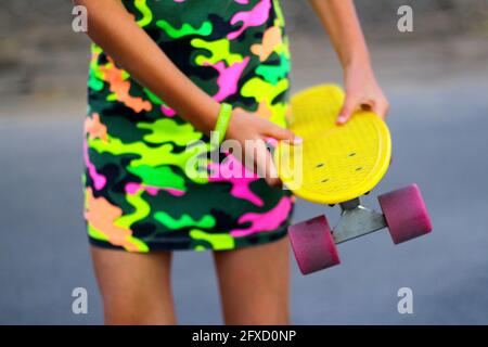 Defokussieren Mädchen mit gelben Penny Board. Vorderansicht. Jugend-Hipster-Kultur. Kinderhände halten tagsüber ein kurzes Cruiser-Pennyboard im Freien. Sporthobby Stockfoto