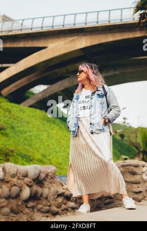 Die durchgehende Hipster-Frau steht mit den Händen auf ihrem jean-Mantel, pinkfarbenen Haaren und einem silbergoldfarbenen Rock im Freien, hinter dem ein Pullover steht. Ind Stockfoto