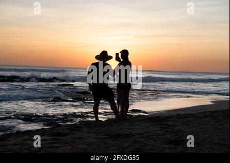 Ein Paar, das mit einem Handy am Strand Fotos vom Sonnenuntergang macht, in einem Paar, das den Sonnenuntergang von einem Strand in Tamarindo, Costa Rica, Mittelamerika, aus beobachtet Stockfoto