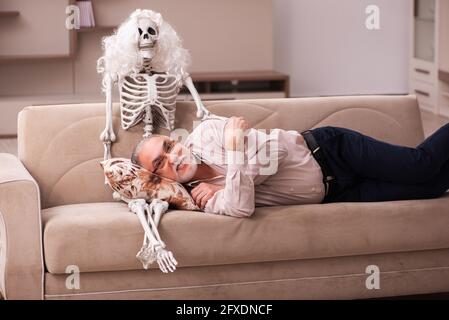 Alter Mann sitzt auf dem Sofa mit dem weiblichen Skelett In unsterbst Liebe Konzept Stockfoto