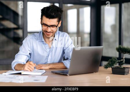 Konzentrierter zufriedener erfolgreicher kaukasischer Geschäftsmann, Freiberufler oder Makler, der eine Brille trägt, an einem Tisch in einem modernen Büro sitzt, einen Laptop benutzt, Notizen macht, Online-Schulungen macht, lächelt Stockfoto