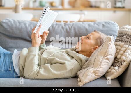 Fröhliche Frau mittleren Alters, die Interessantes Buch liest und sich auf dem Sofa entspannt, Freizeit allein zu Hause verbringt. Selektiver Fokus auf ältere Frauen Stockfoto