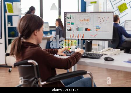 Junge, ungültige, behinderte, gehbehinderte, behinderte Unternehmerin, die mitten im Büro eines Geschäftsgründers auf dem Rollstuhl sitzt und sich auf das multiethnische Team der Kamera in einem Finanzunternehmen konzentriert. Stockfoto