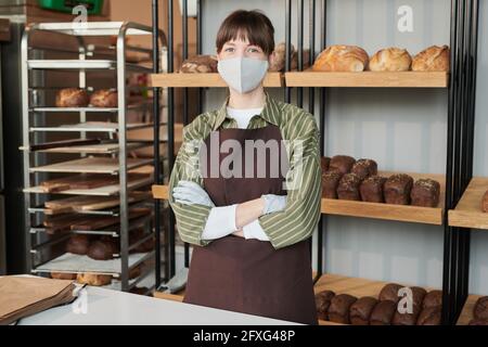 Porträt einer Frau in Uniform und in Maske beim Betragen Kamera, während sie mit gekreuzten Armen in der Bäckerei stand Stockfoto