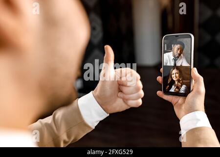 Nahaufnahme eines eleganten Geschäftsmannes, der seinen Kollegen mit dem Daumen die Online-Videokonferenz mit einem modernen Smartphone vorführt. Erfolgreicher Geschäftspartner Stockfoto