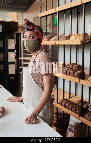 Porträt eines afrikanischen jungen Bäckers in Maske beim Blick auf die Kamera Während der Arbeit in der Bäckerei Stockfoto