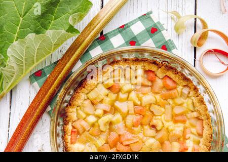 Nahaufnahme der hausgemachten runden gebackenen Rhabarber ( Rheum hybridum ) Pie weiß rustikalen Holz Tisch Hintergrund. Rhabarberstiele an der Seite. Saisonale Küche. Stockfoto