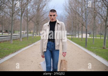Junger Mann im Make-up, der mit Einkaufstaschen spazierengeht und Musik in einem Park hört. Queere Person. Stockfoto