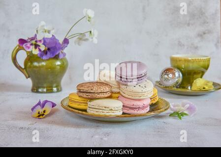 Mehrere bunte Makronen auf einer Keramikuntertasse, eine Tasse Tee und ein Blumenkrug im Hintergrund. Stockfoto