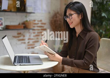 Seitenansicht einer glücklichen Arbeiterin, die während des Schreibens auf einem Notizbuch schrieb Arbeiten mit Laptop im Café Stockfoto